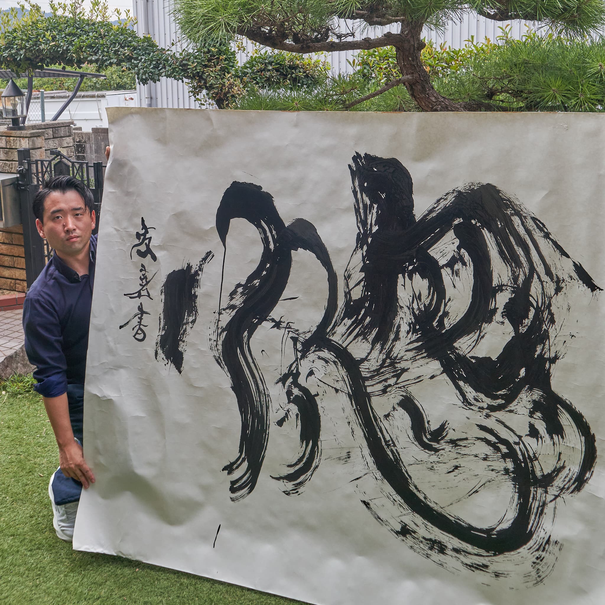 屋外で大判の紙に書いた大字「惚」を、作者が広げて見せている