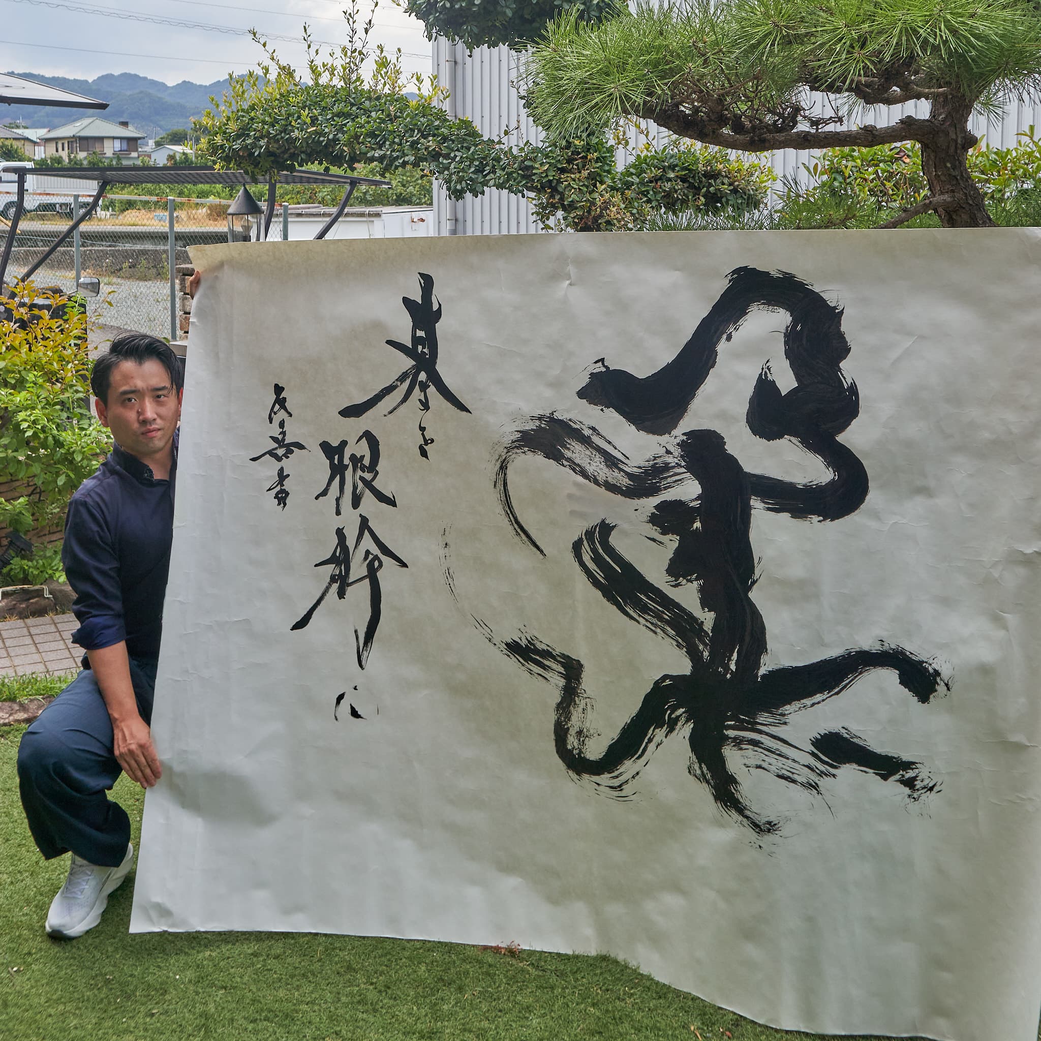 屋外で大判の紙に書いた大字「柔」を、作者が広げて見せている。左に細字の詞を添える