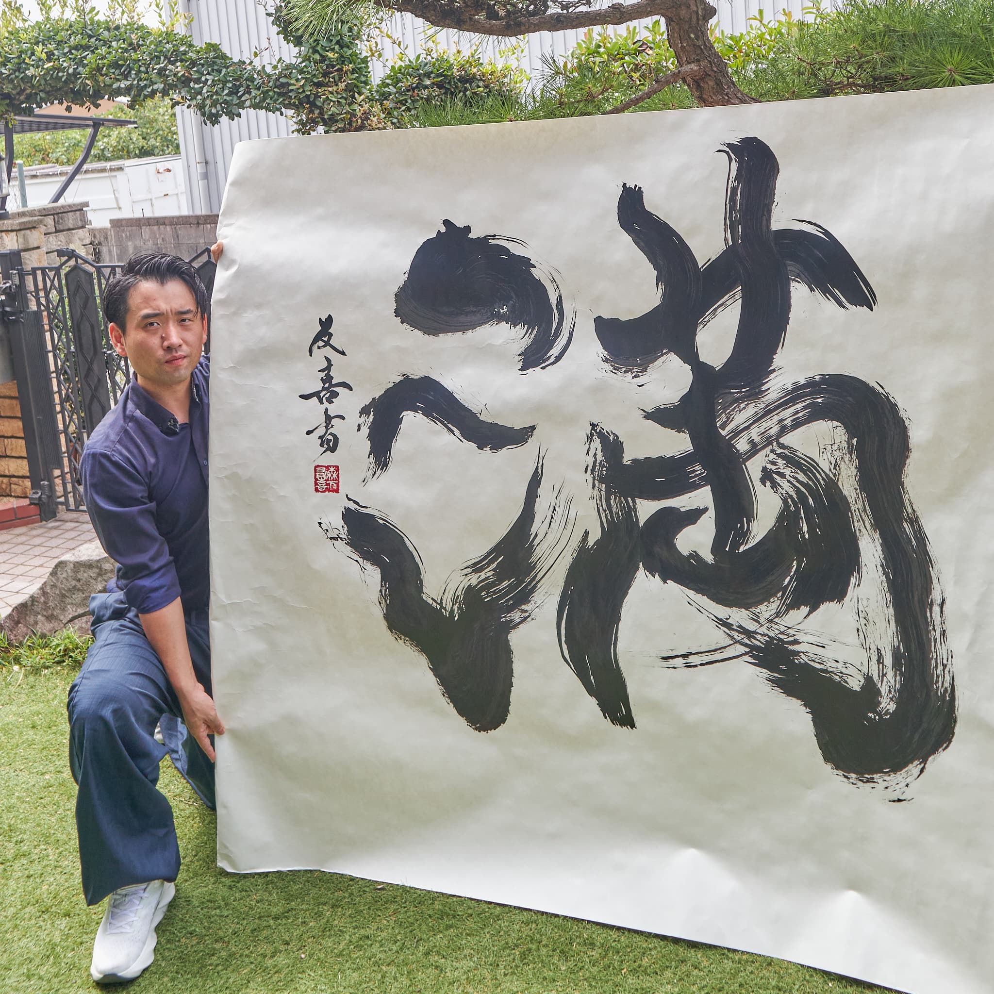 屋外で大判の紙に書いた大字「満」を、作者が広げて見せている