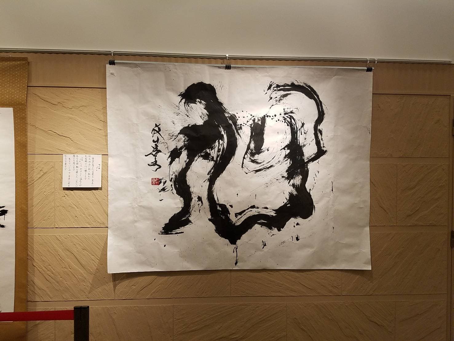 展示会場の壁に掛けた大型の墨書「心」。傍らに作品解説の札を添えている