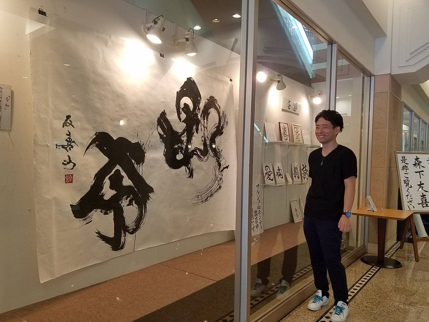 個展会場の入口に立つ作者と、壁面に掛けた大型作品