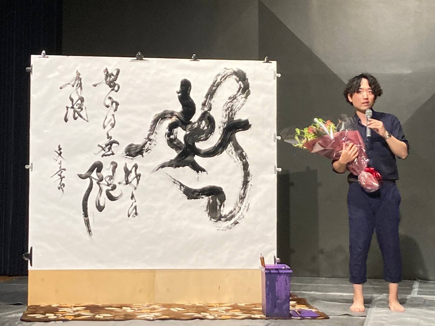 舞台上の大型作品の傍らで、花束を手にマイクを持つ作者